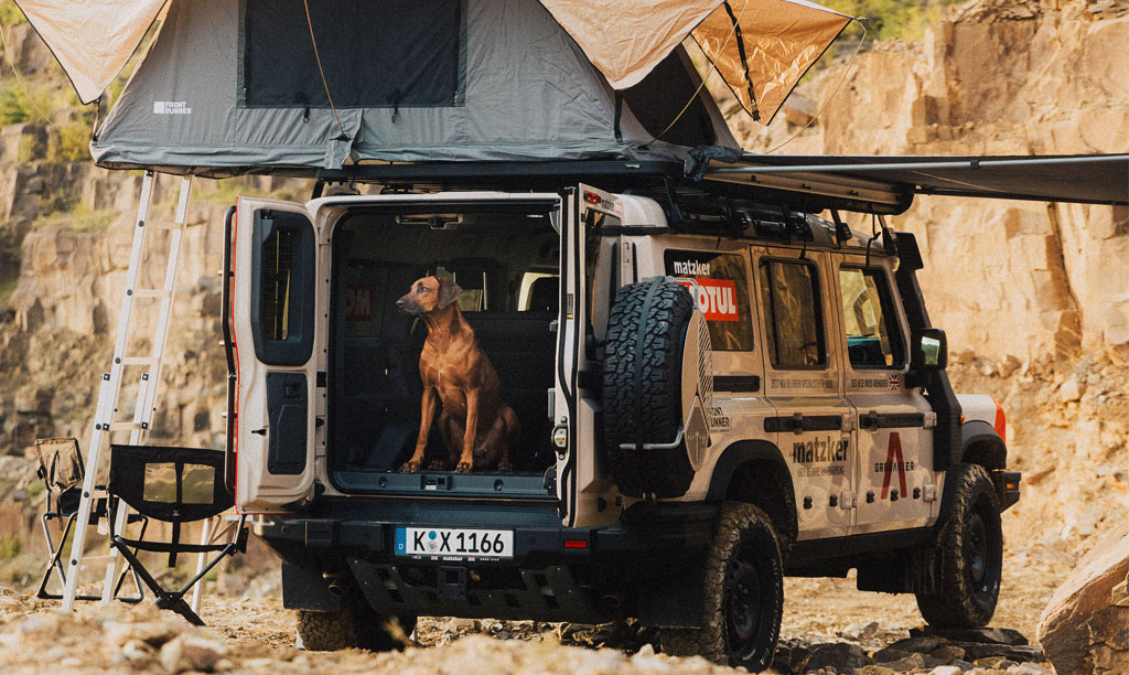 Ein Offroad-Fahrzeug mit einem Dachzelt, das in einer felsigen Umgebung parkt. Ein Hund sitzt im offenen Fahrzeugheck, bereit für das Abenteuer.
