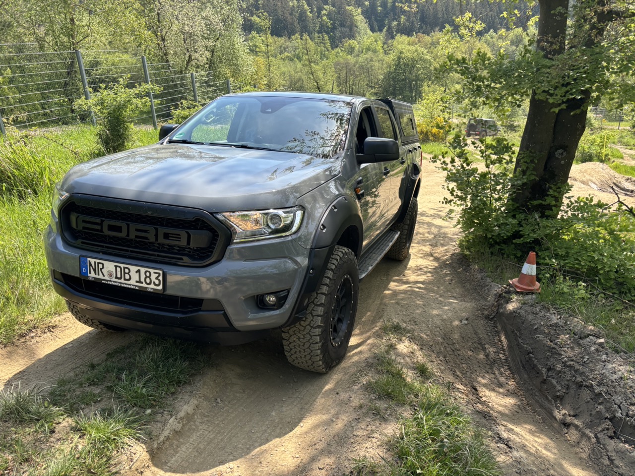 Ein grauer Ford Ranger steht auf einem unbefestigten Weg, umgeben von grünem Gras und Bäumen, mit einem Verkehrshütchen im Vordergrund.