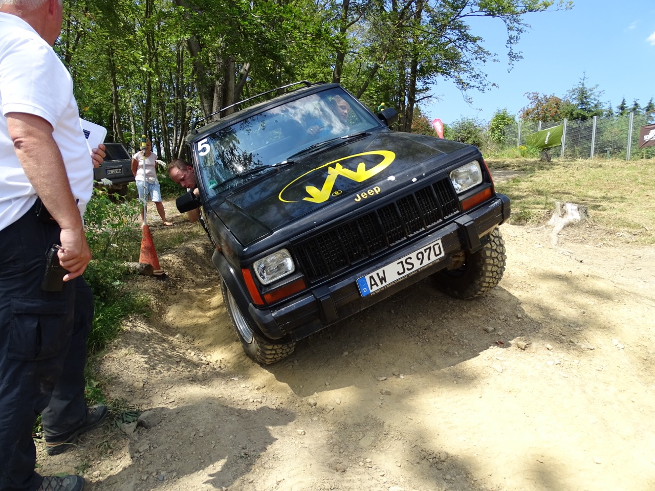 Ein schwarzer Jeep mit gelbem Logo in einer leichten Neigung auf einem unebenem Weg, während Zuschauer anwesend sind.