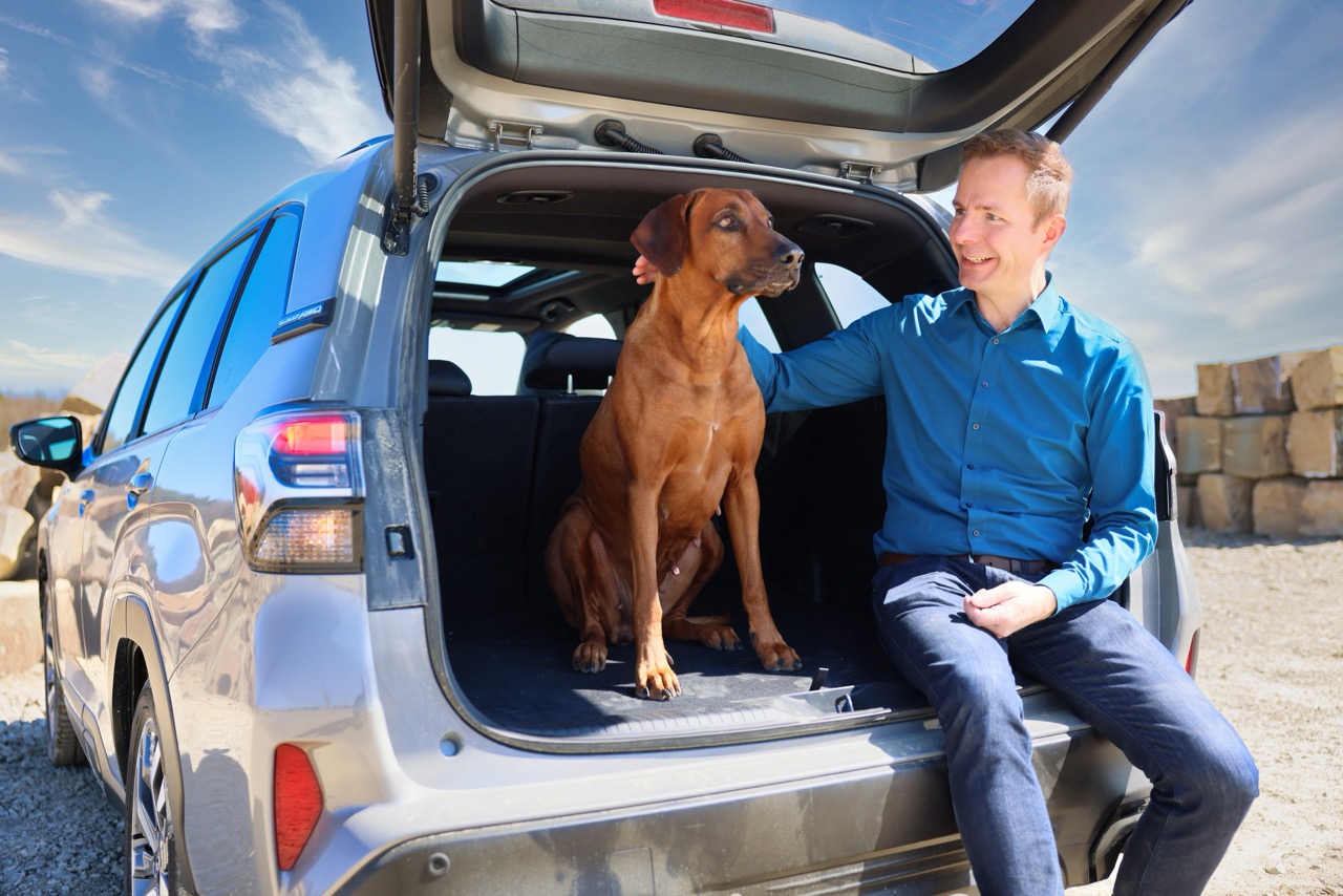 Mann sitzt im Kofferraum seines Autos und streichelt einen Hund bei sonnigem Wetter.