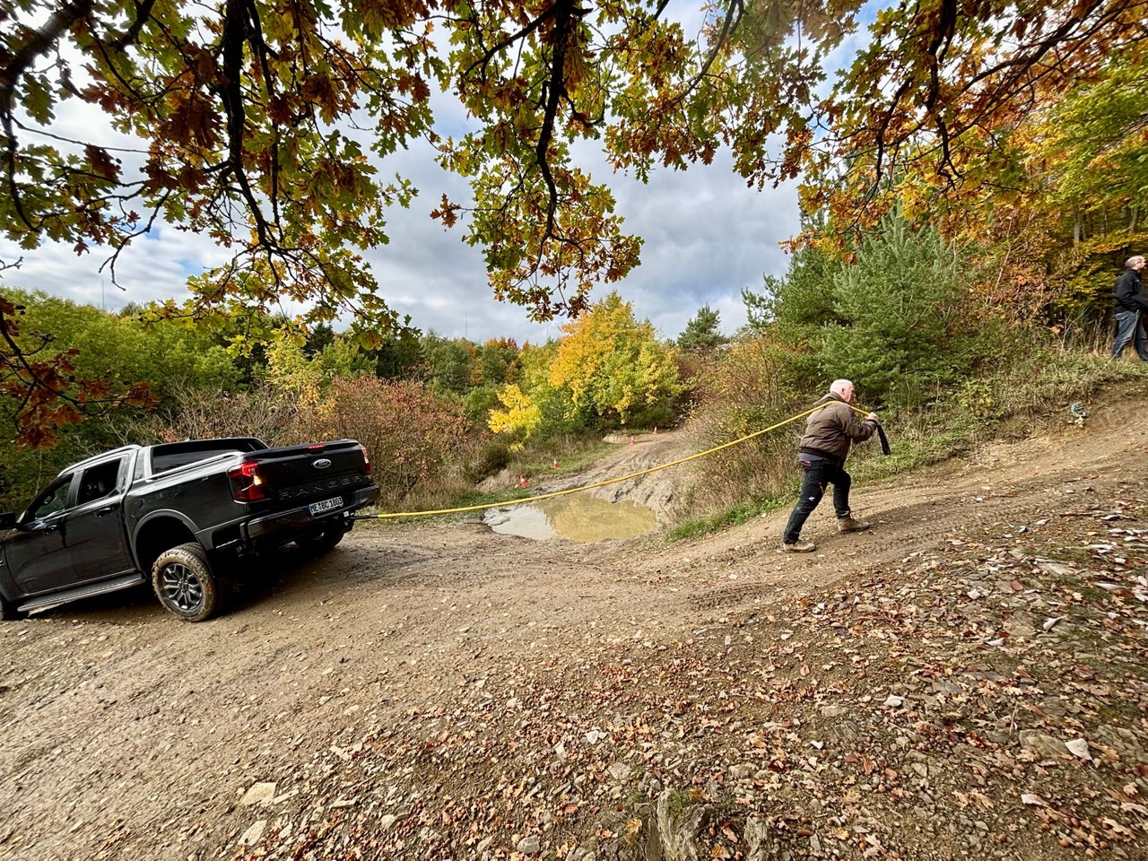 Ein Mann zieht mit einem Seil einen Geländewagen auf einem unebenen, schmutzigen Weg in einer herbstlichen Umgebung.