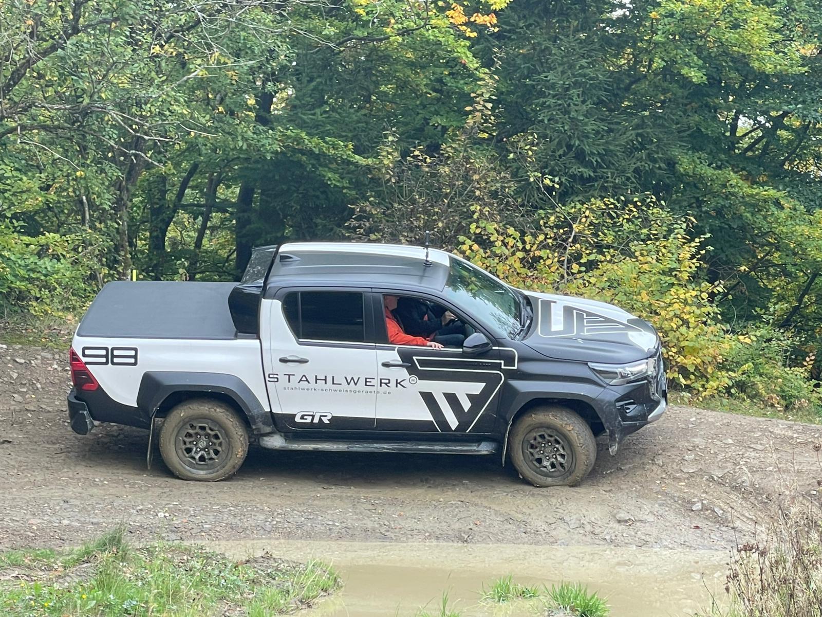 Ein SUV mit Stahlwerk-Design fährt auf einem unbefestigten Weg in einer Waldumgebung.
